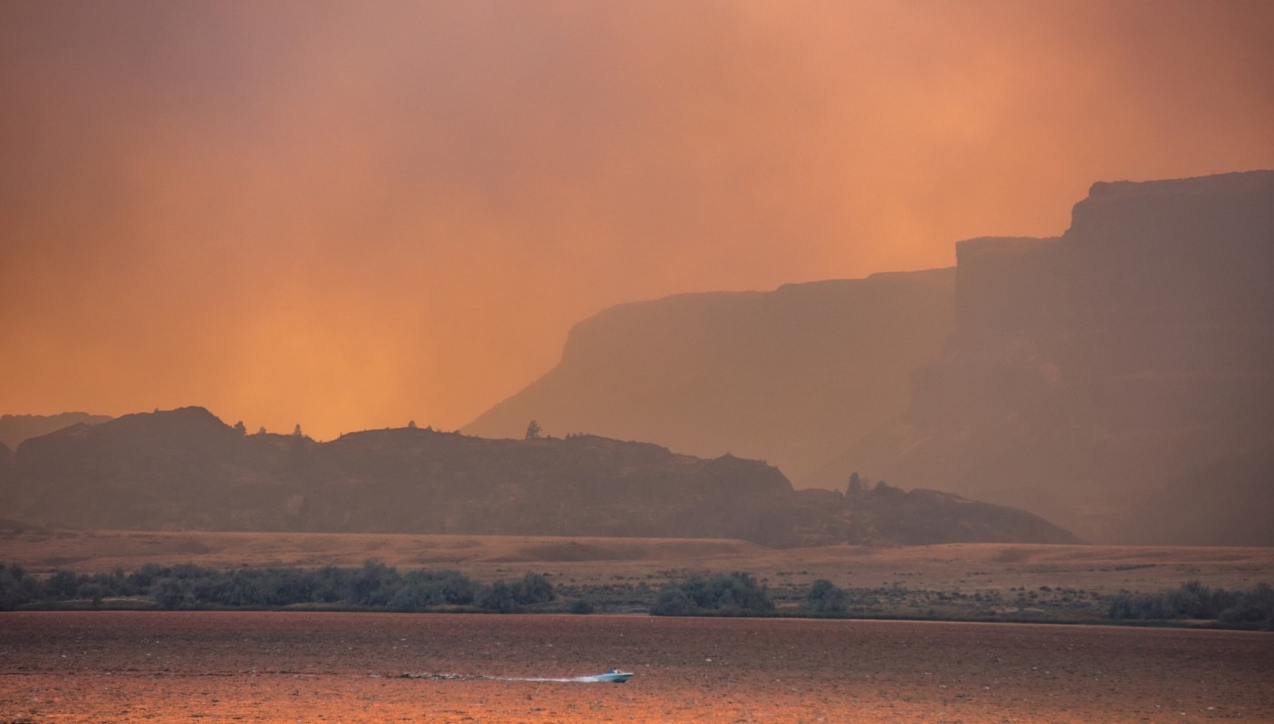 A boat moves across a lake with smoke-filled skies and silhouettes of mountains in the background.