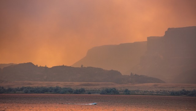A boat moves across a lake with smoke-filled skies and silhouettes of mountains in the background.