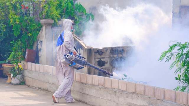 A person in protective gear operates a fogging machine in a garden.