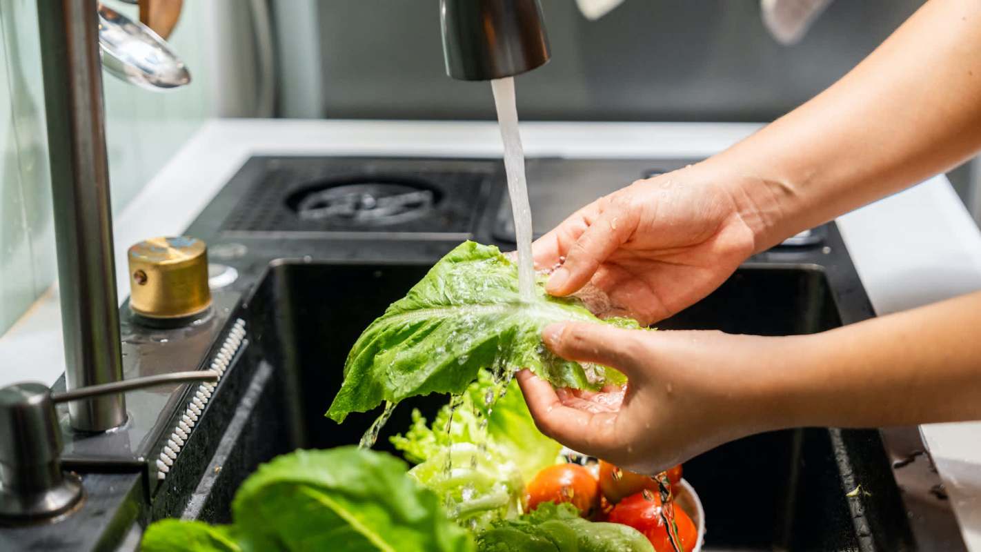 A person washes lettuce under running water in a sink filled with vegetables.