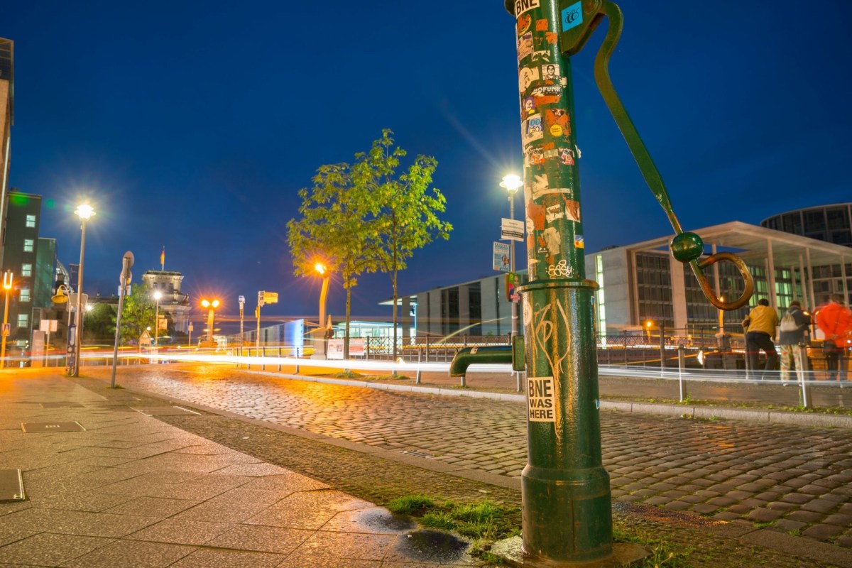 A green water hand pump on the sidewalk along a road in Berlin Germany