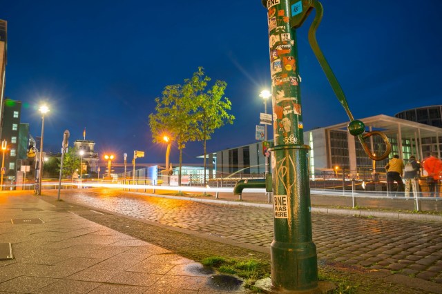 A green water hand pump on the sidewalk along a road in Berlin Germany