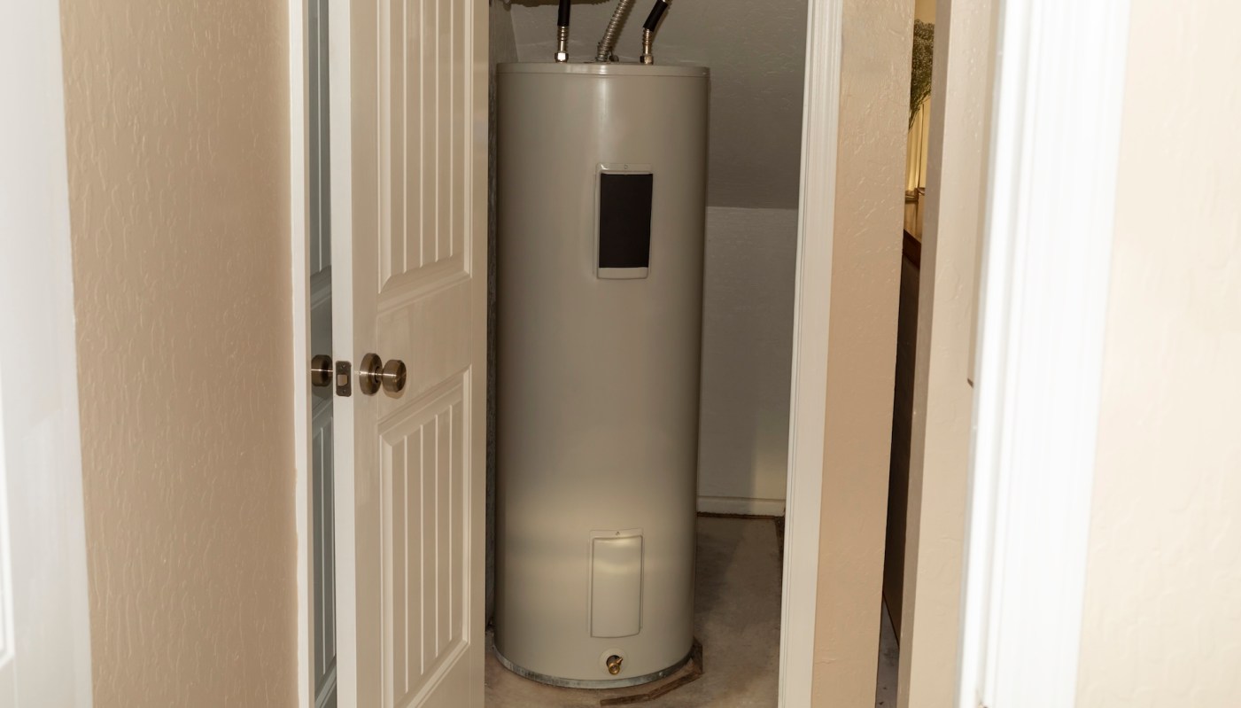 A water heater in a small, enclosed space with a door partially open.