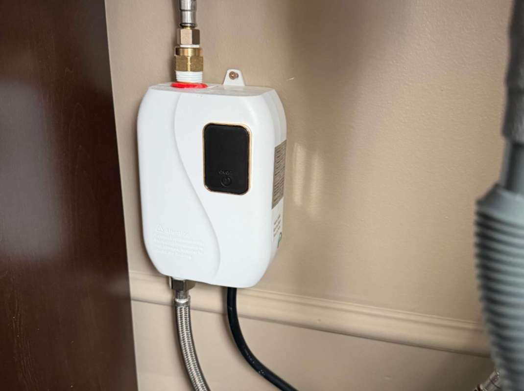A white device mounted on a wall, connected to pipes, with a black button and warning label.