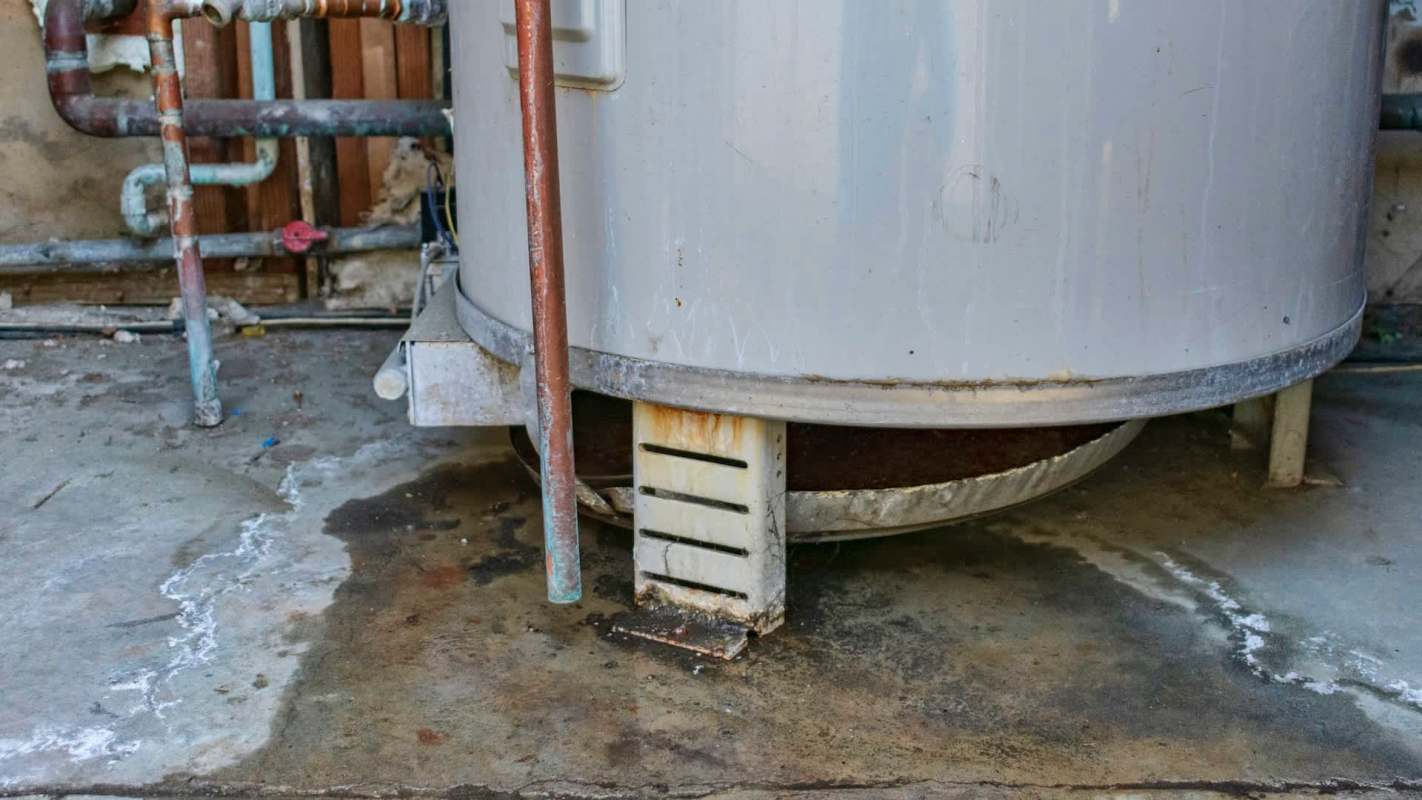A large, rusted water heater sits on a concrete floor with visible water stains around it.