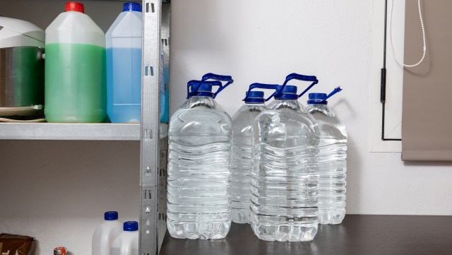 Four large bottles of water with blue caps are on a shelf next to various cleaning liquids.