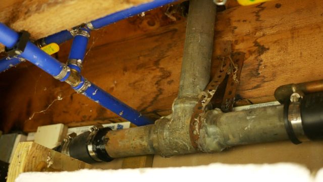 Pipes and plumbing components are visible beneath a wooden surface with some cobwebs.