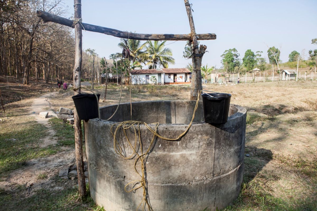 A wooden well with buckets stands in a field near a house and trees.