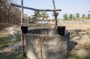 A wooden well with buckets stands in a field near a house and trees.