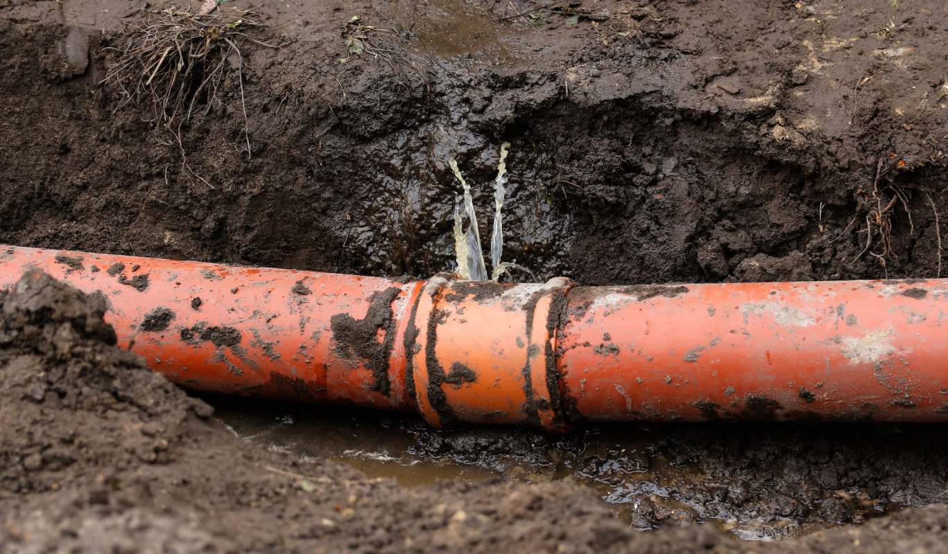 An orange pipe leaking water in a trench surrounded by dirt.