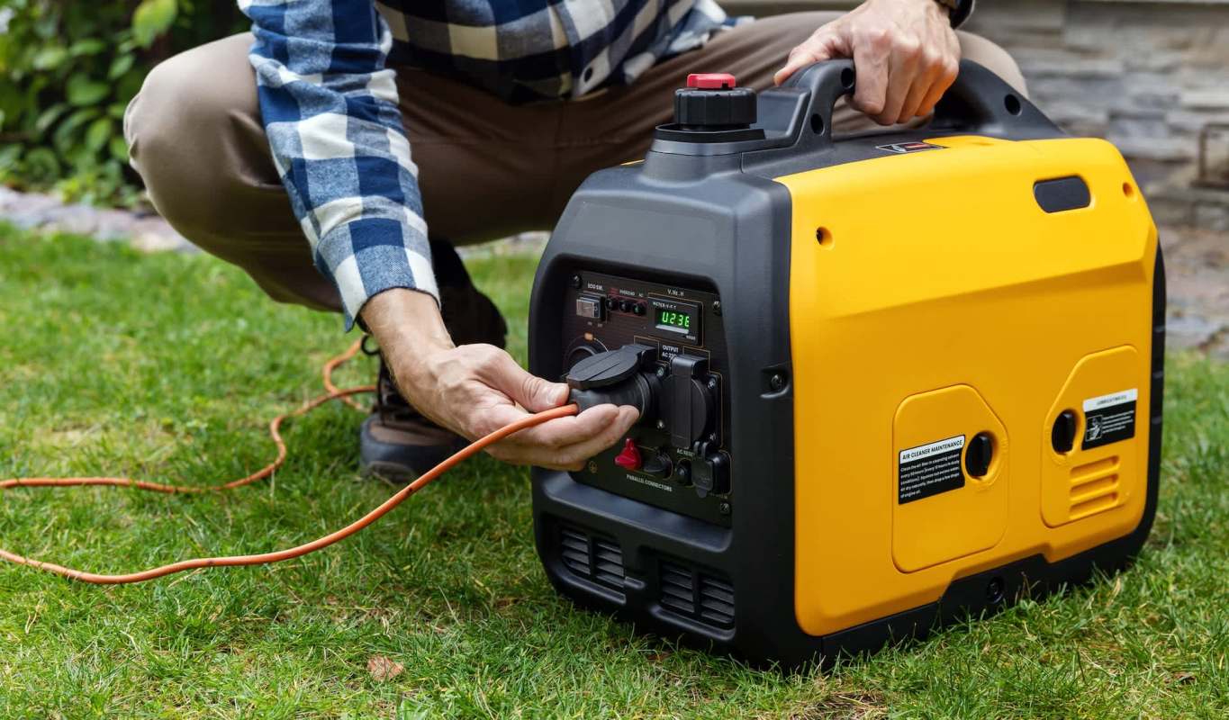 A person plugs an orange extension cord into a portable generator on the grass.