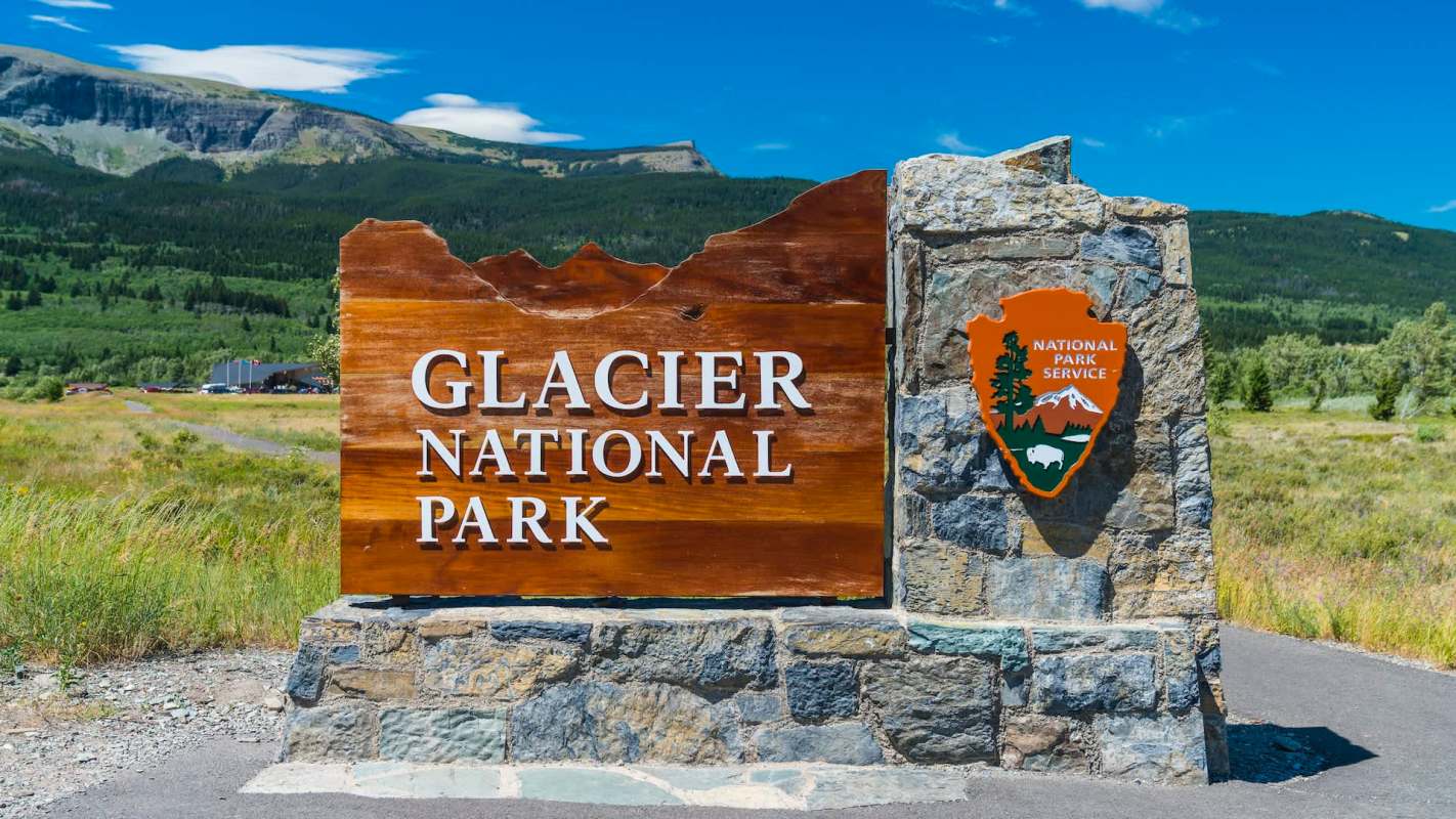 A sign for Glacier National Park with a stone base and a National Park Service emblem in a scenic outdoor setting.