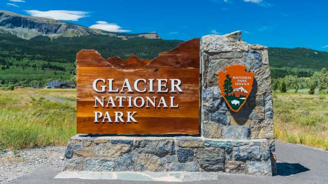 A sign for Glacier National Park with a stone base and a National Park Service emblem in a scenic outdoor setting.