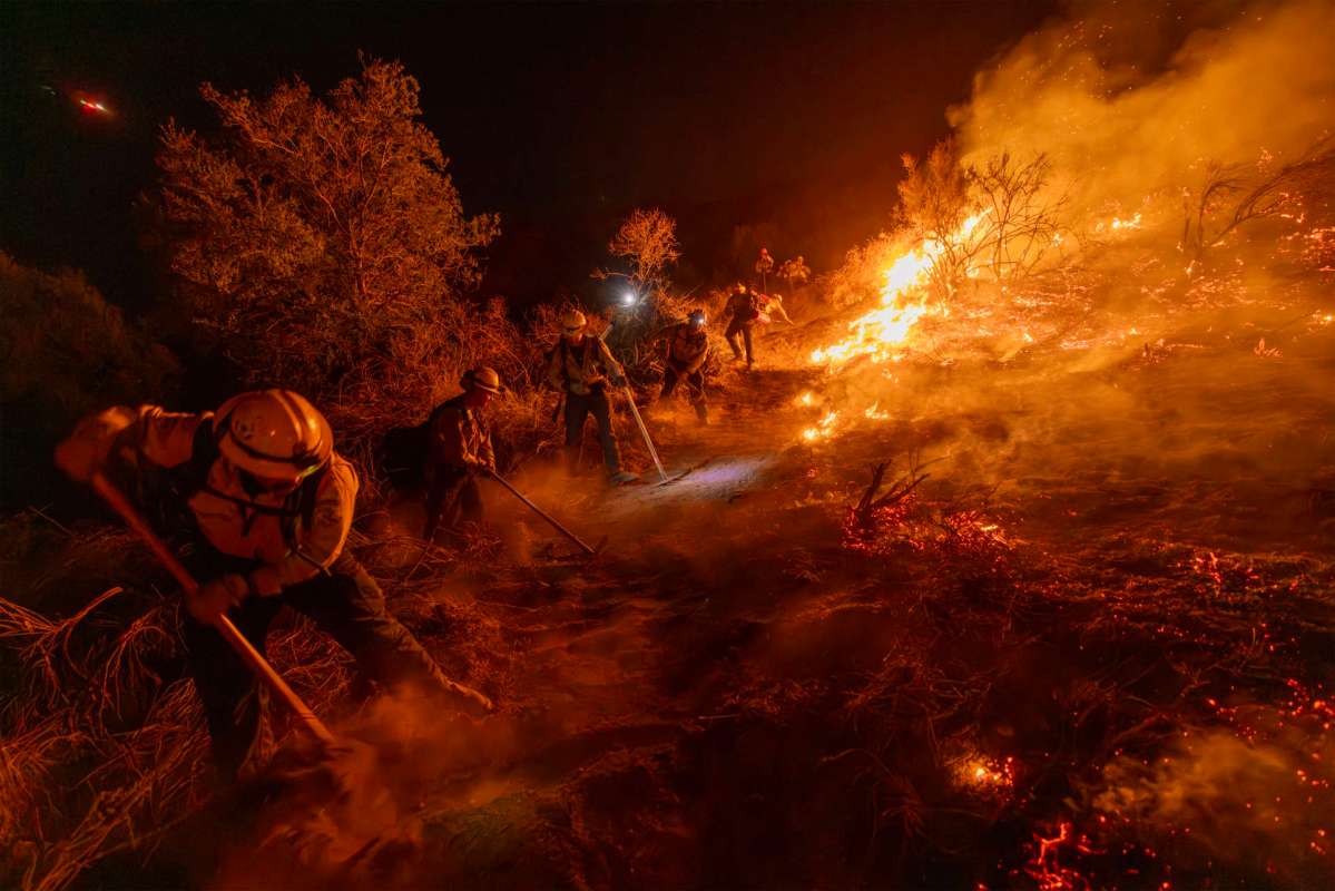 Firefighters work to contain a wildfire at night, using tools to combat flames and smoke in a rugged terrain.