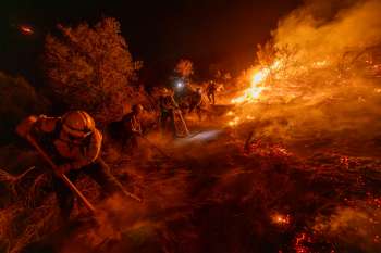 Firefighters work to contain a wildfire at night, using tools to combat flames and smoke in a rugged terrain.
