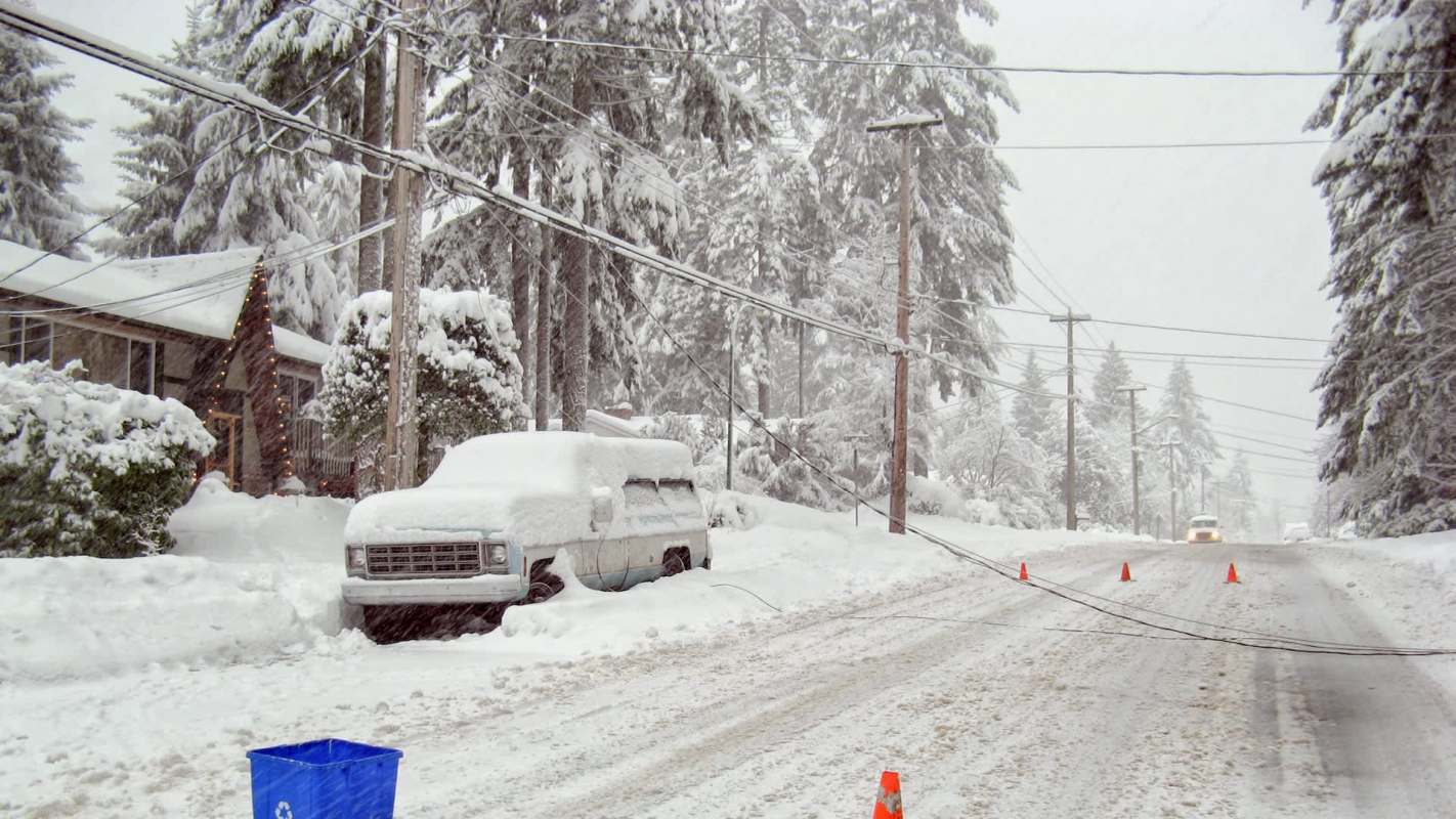 A snowy street with a covered vehicle, downed power lines, and a recycling bin and orange cones in the foreground.